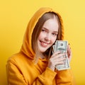 Emotional happy teenage blonde girl in hood win money cash holding dollars in hands isolated on color yellow background. Portrait