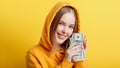 Emotional happy teenage blonde girl in hood win money cash holding dollars isolated on color yellow background. Portrait