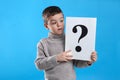 Emotional little boy holding paper with question mark on light blue background Royalty Free Stock Photo