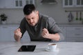 Emotional man with tablet at table in kitchen. Online hate concept