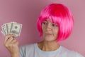 Portrait of a young beautiful confused woman with pink hair holding a wad of dollars on a pink background Royalty Free Stock Photo