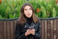 Emotional portrait of a young pretty frustrated brunette woman sitting outdoors city park wearing black leather coat using smartph