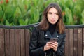 Emotional portrait of a young pretty frustrated brunette woman sitting outdoors city park wearing black leather coat using smartph