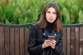 Emotional portrait of a young pretty frustrated brunette woman sitting outdoors city park wearing black leather coat using smartph