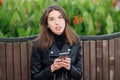 Emotional portrait of a young pretty frustrated brunette woman sitting outdoors city park wearing black leather coat using smartph