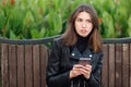 Emotional portrait of a young pretty frustrated brunette woman sitting outdoors city park wearing black leather coat using smartph