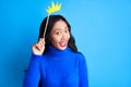 Emotional suprised woman with paper crown on stick on blue background