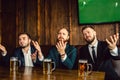 Emotional and upset young men in suits sit at table in bar. They wave with hands and look forward. Guys watch football