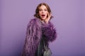 Emotional white girl with colorful makeup posing on purple background with purse. Indoor photo of amazed curly woman in