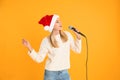 Emotional woman in Santa Claus hat singing with microphone on yellow. Christmas music Royalty Free Stock Photo