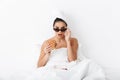 Emotional woman with towel on head lies in bed under blanket isolated over white wall background wearing sunglasses eat burger