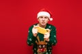 Emotional young gamer man in hat with santa hat and warm sweater stands with smartphone in hand on red background and looks at