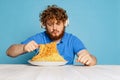 Emotional young hairy red-bearded man tasting large portion of noodles, pasta isolated on blue studio background.