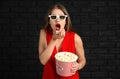 Emotional young woman with cup of popcorn and 3D cinema glasses against dark brick wall Royalty Free Stock Photo