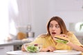 Emotional young woman eating sandwich instead of salad in kitchen