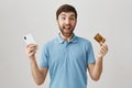Emotive positive adult bearded boyfriend holding cellphone and credit card, standing with cheerful expression over gray
