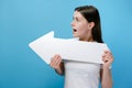 Emotive young brunette woman with shocked face holding white paper arrow and points sideways, dressed in t-shirt