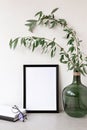 Empty black picture frame mockup, green glass vase with tree branch, stationery on beige tabletop and wall background Royalty Free Stock Photo