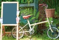 An empty blackboard on wooden stand ready for put text in, besides of its is beautiful foldable bike, background is green plant