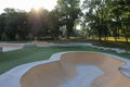 Empty concrete skateboard park at sunrise. Playground in the background, with rays of sunrise shinging through trees Royalty Free Stock Photo