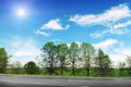 An empty countryside road with trees and bushes against a blue sky with clouds and sun Royalty Free Stock Photo