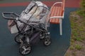 Empty four-wheeled black pram covered with anti-rain cellophane canopy, standing next to brown bench on playground on rainy day