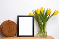 Empty frame standing on small wooden table with yellow tulips and straw bag Royalty Free Stock Photo