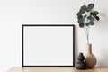 Empty horizontal frame mockup in modern minimalist interior with plant in trendy vase on white wall