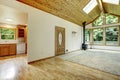 Empty living room with connected kitchen, vaulted wood ceiling, and large windows.