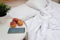Empty messy unmade bed with white pillows and blanket, bedside table with peaches, mobile phone and plant in pot Royalty Free Stock Photo