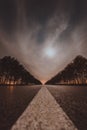 Empty night asphalt road, bright full moon behind clouds and city light behind two rows of trees on both side of road