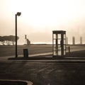 An empty parking lot with a street lamp, phone booth and trash can on a foggy morning with water and shadows in the background