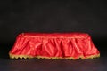 Empty pedestal covered with bright red silk carpet, golden fringe on black background.