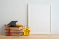 Empty photo frame, textbooks, graducation hat, pencil and alarm clock on wooden table with white wall background copy space.