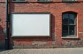 Empty rectangular billboard on rustic red brick wall. Nearby is old window, drainpipe. Paved sidewalk in foreground. Offers blank Royalty Free Stock Photo