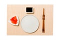 Empty round white plate with chopsticks for sushi, ginger and soy sauce on brown sushi mat background. Top view with copy space