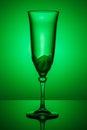 Empty transparent champagne glass on a thin high leg with reflection on a dark glossy surface in green backlight