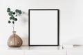 Empty vertical frame mockup in modern minimalist interior with plant in trendy vase on white wall background.