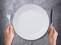 Empty white ceramic plate with hand with knife and fork on dark table gray cement background