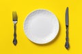 Empty white dinner plate, table knife and fork against yellow background. Blank mockup template for unbalanced diets and poor Royalty Free Stock Photo