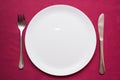 Empty white plate, fork and knife on maroon tablecloth. Elegant table setting. Top view, closeup Royalty Free Stock Photo