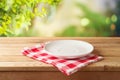 Empty white plate on wooden table with red checked tablecloth over  blurred nature background.  Kitchen mock up for design and Royalty Free Stock Photo