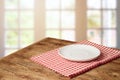 Empty white plate on wooden table with red checked tablecloth over  blurred window background.  Kitchen mock up for design and Royalty Free Stock Photo