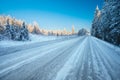 Empty winter road with ice-slick and snow through snowy fields and forest.