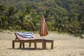 Empty wooden beach loungers with mattresses and a folded sun umbrella on the white sand against the backdrop of the green palm Royalty Free Stock Photo