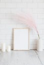 Empty wooden picture frame mockup, candles, pink dried flowers in vase on table. White tiles wall background. Minimal, elegant,