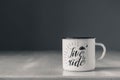 Enamel white metal mug with black line on the edge with the inscription on white wooden table on gray background. Place for text