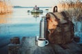 Mug of coffee or tea, backpack of traveller and thermos on wooden pier on tranquil lake. A fisherman on rubber boat. Royalty Free Stock Photo