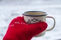 Enameled mug with hot tea in a hand in a red warm mitten in winter on a snowy background. Royalty Free Stock Photo