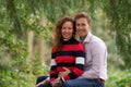 Enamored couple sits on a thick tree trunk, the man sits behind the woman. looking at the camera